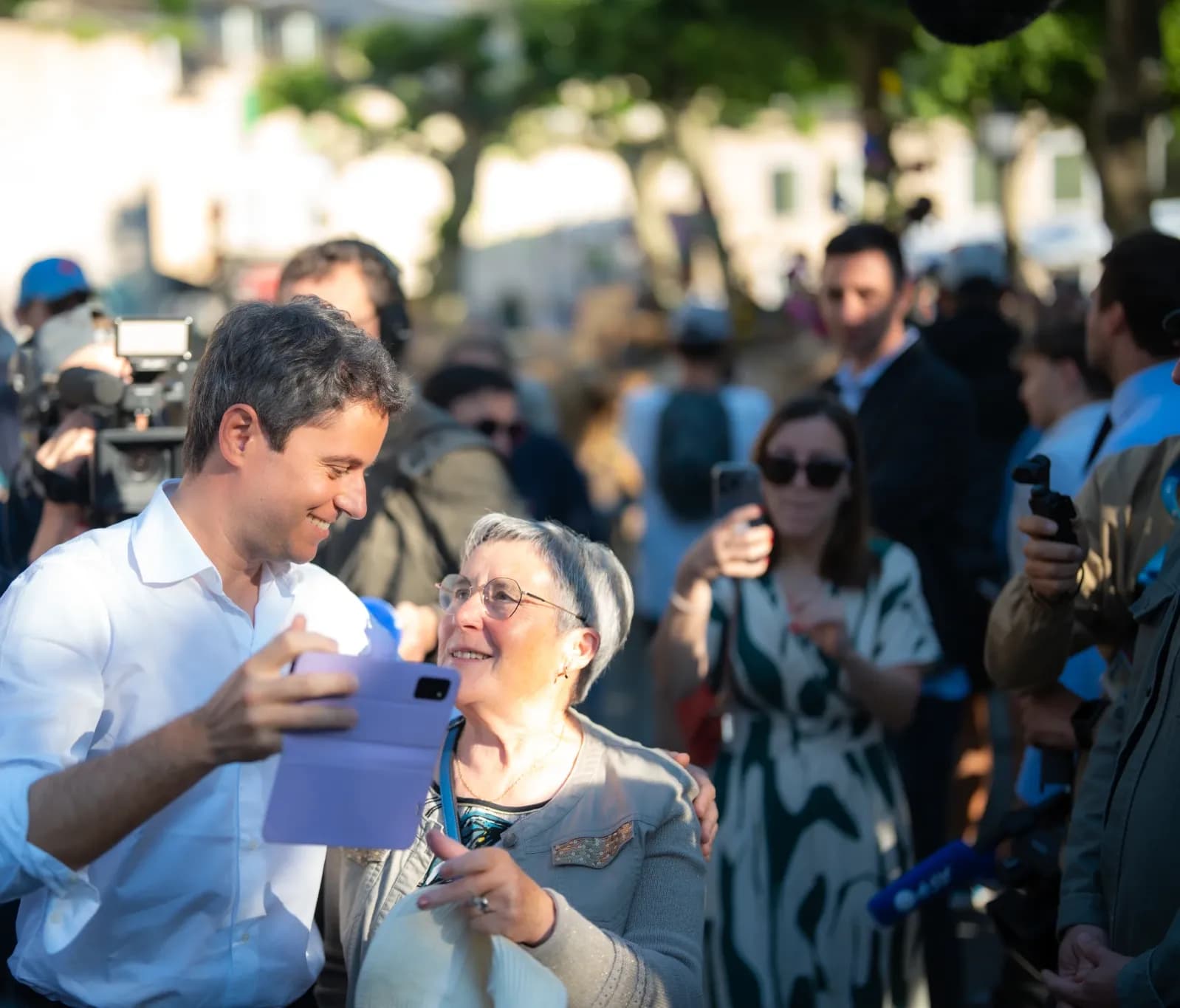 Gabriel Attal, en chemise blanche, sourit et tend un téléphone pour un selfie avec une habitante aux cheveux gris, sur une place arborée, au milieu de la foule.