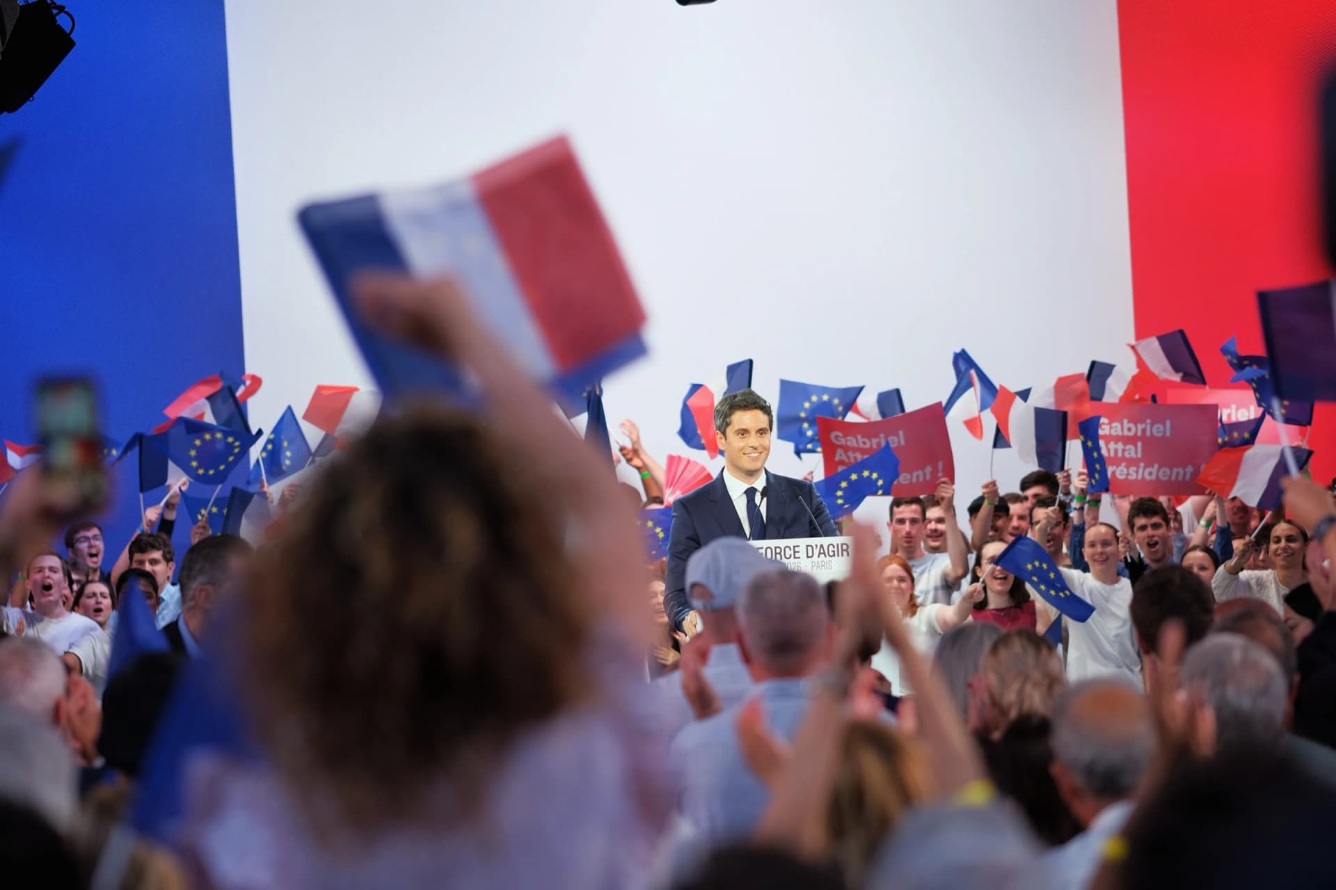 Meeting de campagne : Gabriel Attal à la tribune, devant une salle qui agite des drapeaux français et européens.