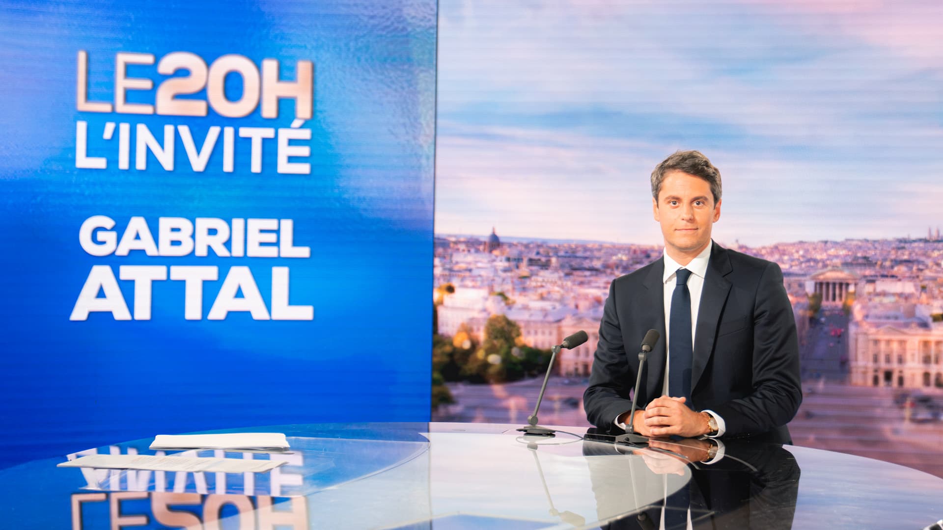 Photographie sur le plateau du journal télévisé de TF1. Gabriel Attal, en costume sombre et cravate bleue, est assis derrière le bureau circulaire réfléchissant du JT, les mains croisées devant lui, regardant face à l'objectif. Deux microphones sont posés sur le bureau. À sa gauche, un grand écran bleu affiche en lettres blanches : « LE 20H – L'INVITÉ – GABRIEL ATTAL ». En arrière-plan, un décor représentant une vue panoramique de Paris aux tons chauds du soir.