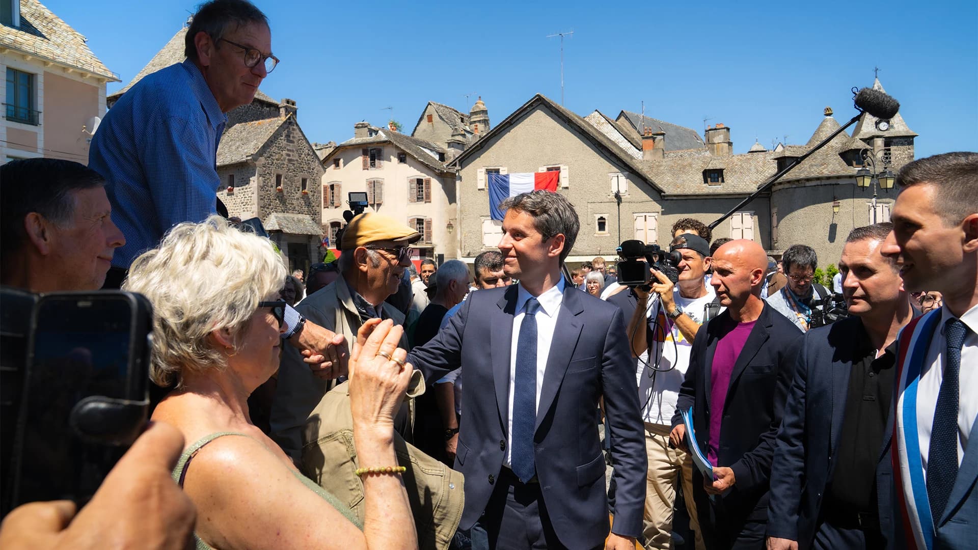 Photographie en extérieur par temps ensoleillé sur une place de village aux bâtisses en pierre grise. Au centre, Gabriel Attal, en costume bleu marine et cravate sombre, avance en souriant au milieu d'une foule. Autour de lui, des habitants l'accueillent chaleureusement — une femme aux cheveux blancs lui tend la main au premier plan — tandis que plusieurs caméramans et photographes captent la scène. À droite, un élu local portant une écharpe tricolore bleu-blanc-rouge l'accompagne. En arrière-plan, un drapeau français est accroché à la façade d'un bâtiment, sous un ciel bleu sans nuages.