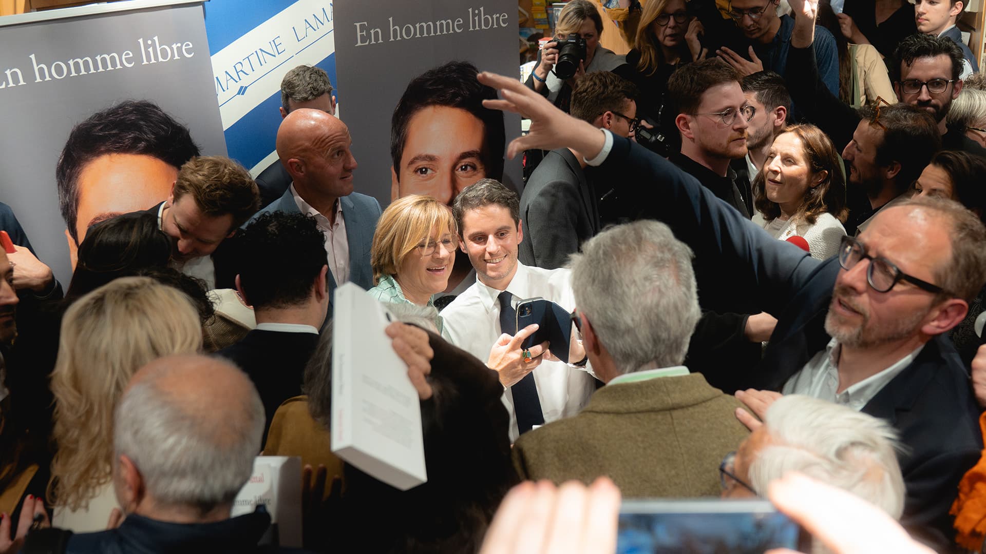 Photographie d'une séance de dédicace de Gabriel Attal dans une librairie bondée. Au centre de l'image, Gabriel Attal en chemise blanche et cravate sombre sourit en prenant un selfie avec une femme à lunettes aux cheveux blonds. Il tient un stylo à la main, vraisemblablement en train de signer des livres. Autour d'eux, une foule dense de personnes, dont plusieurs photographes et caméramans. En arrière-plan, deux bannières verticales reprennent la couverture du livre En homme libre et l'enseigne de la librairie Martine Lamartine.