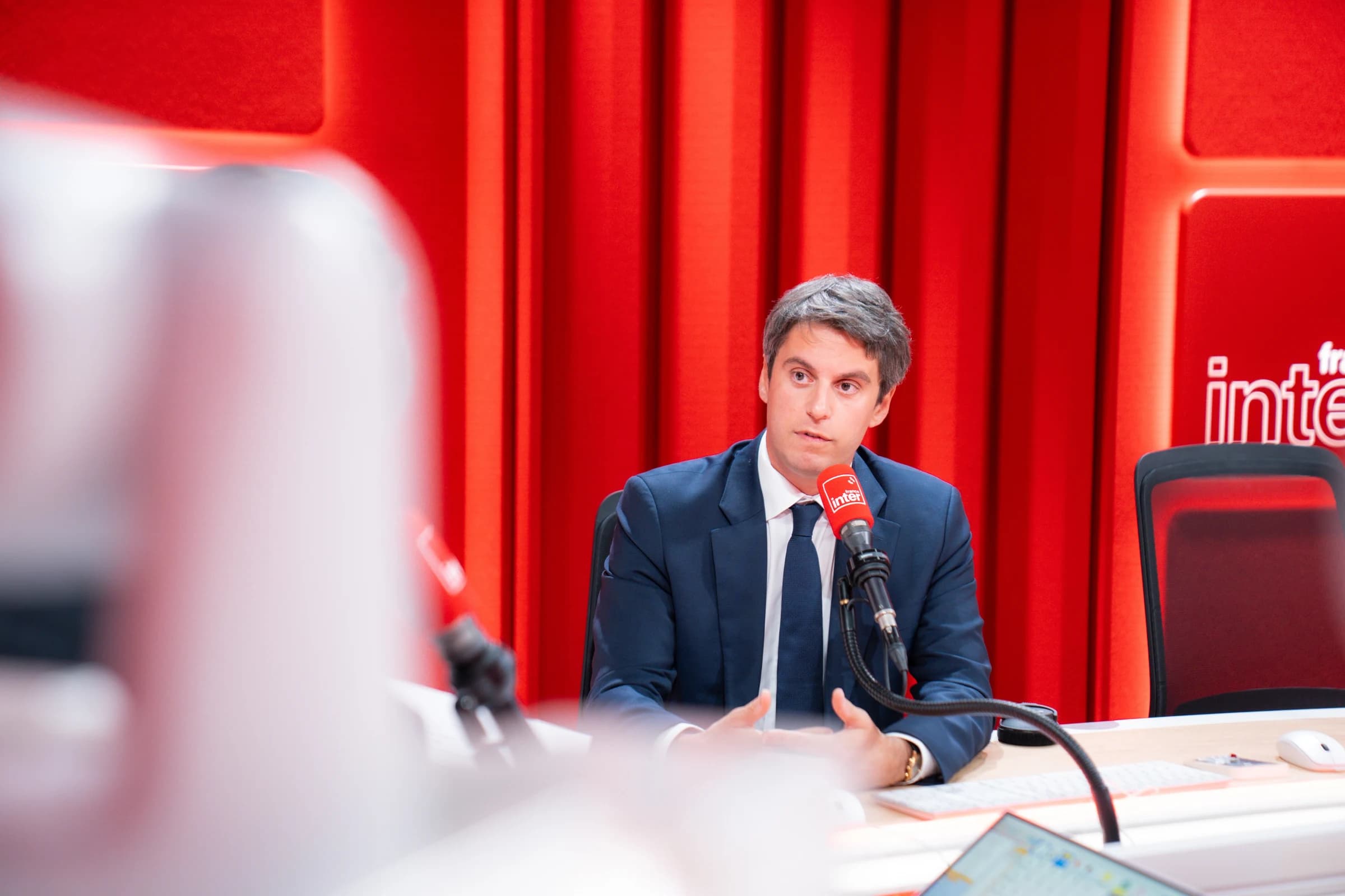 Gabriel Attal en costume bleu marine, assis dans un studio de France Inter, parlant devant un micro rouge de la station, vu depuis le fond de la pièce avec du matériel technique au premier plan.