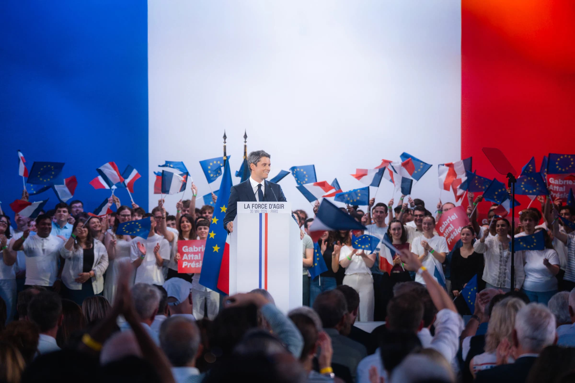 Photographie de meeting politique. Gabriel Attal, en costume sombre et cravate noire, se tient debout derrière un pupitre blanc orné d'une bande tricolore et portant l'inscription « LA FORCE D'AGIR – 30 MAI 2026 – PARIS ». Il sourit en regardant légèrement sur le côté. Derrière lui, un immense drapeau français bleu-blanc-rouge occupe toute la scène en arrière-plan. Une foule de jeunes supporters vêtus de blanc l'entoure, brandissant des drapeaux français et européens ainsi que des pancartes rouges « Gabriel Attal Président ! ». Au premier plan, le public, de dos, applaudit. L'atmosphère est festive et très mobilisée.