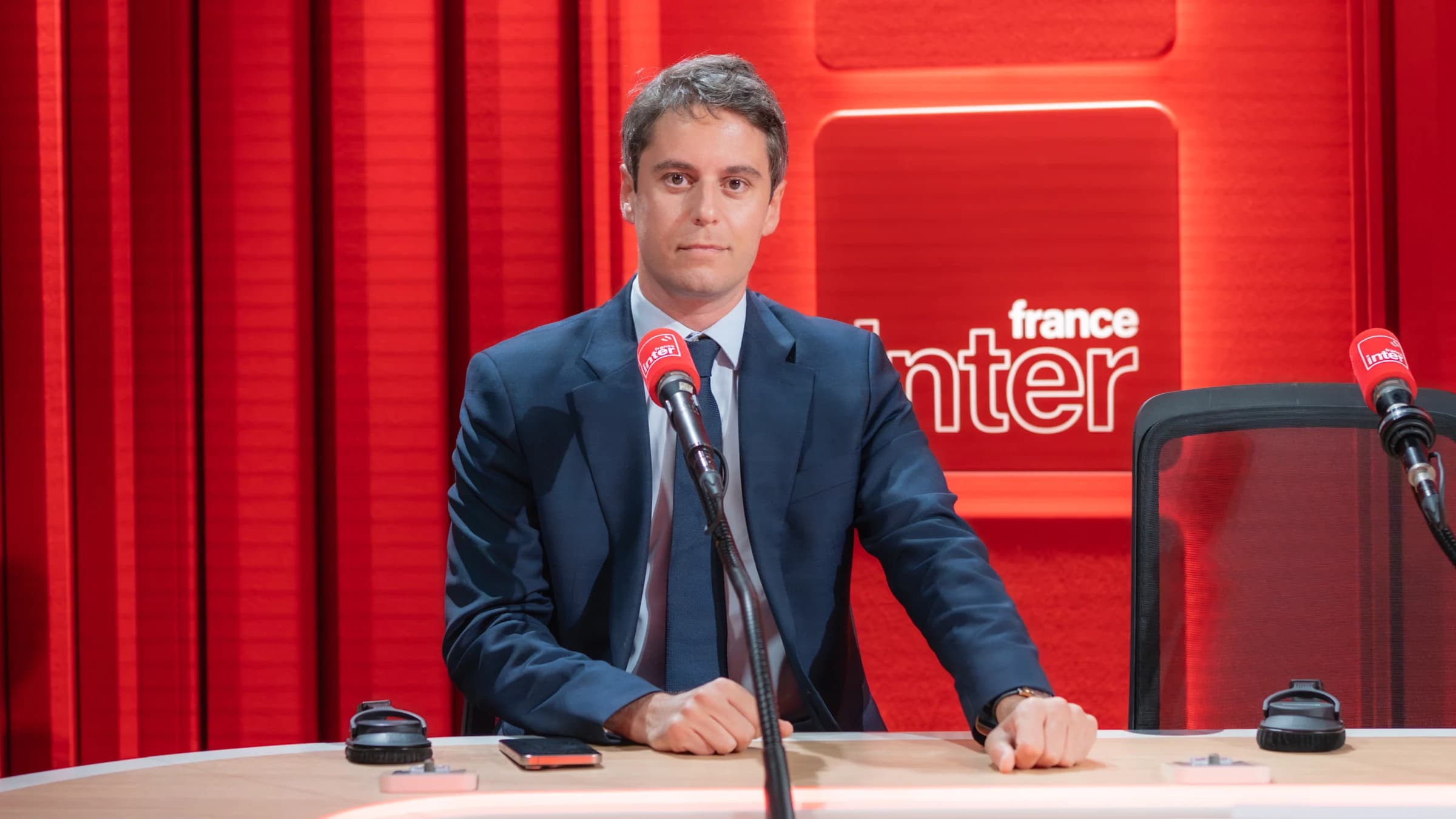 Portrait de Gabriel Attal en costume sombre, assis face caméra à une table de studio devant un micro rouge, avec le logo de France Inter en arrière-plan.