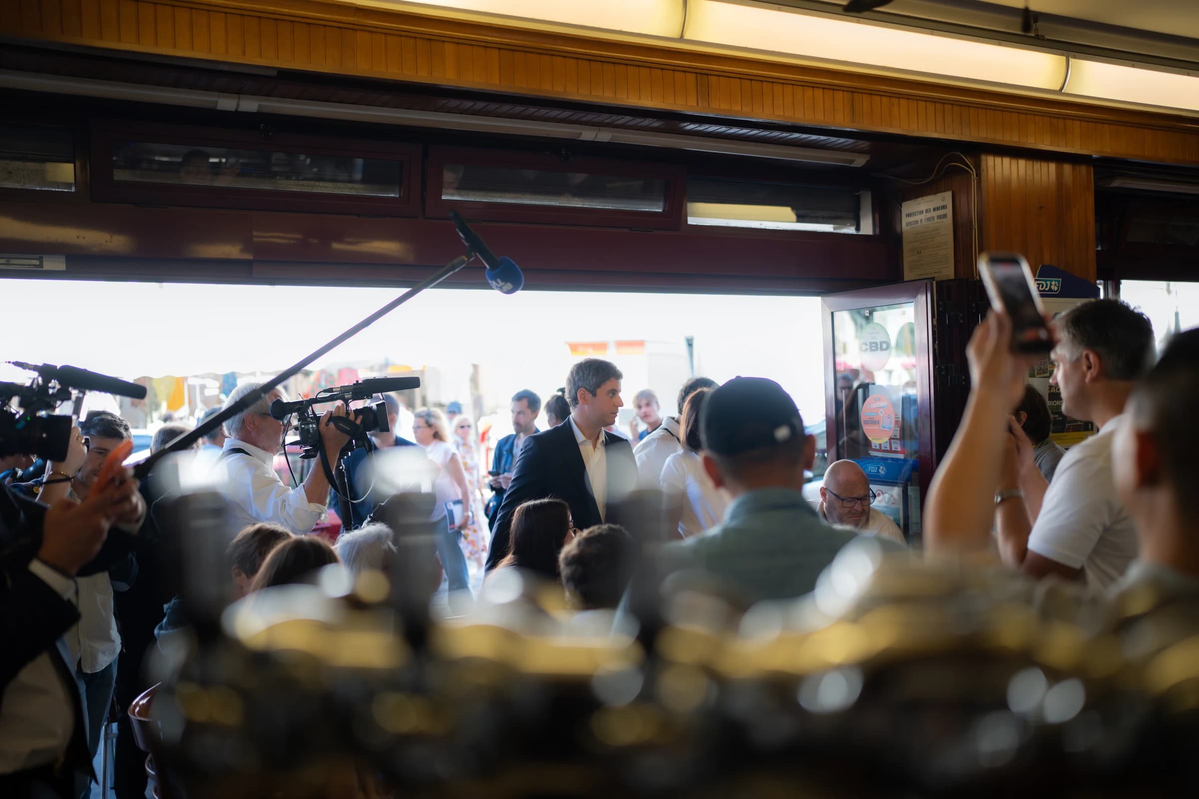 Gabriel Attal en déplacement à Villers-Cotterêts, photographié depuis l'intérieur d'un café. Il avance au milieu d'une foule compacte, encerclé par des caméras de journalistes et des smartphones.