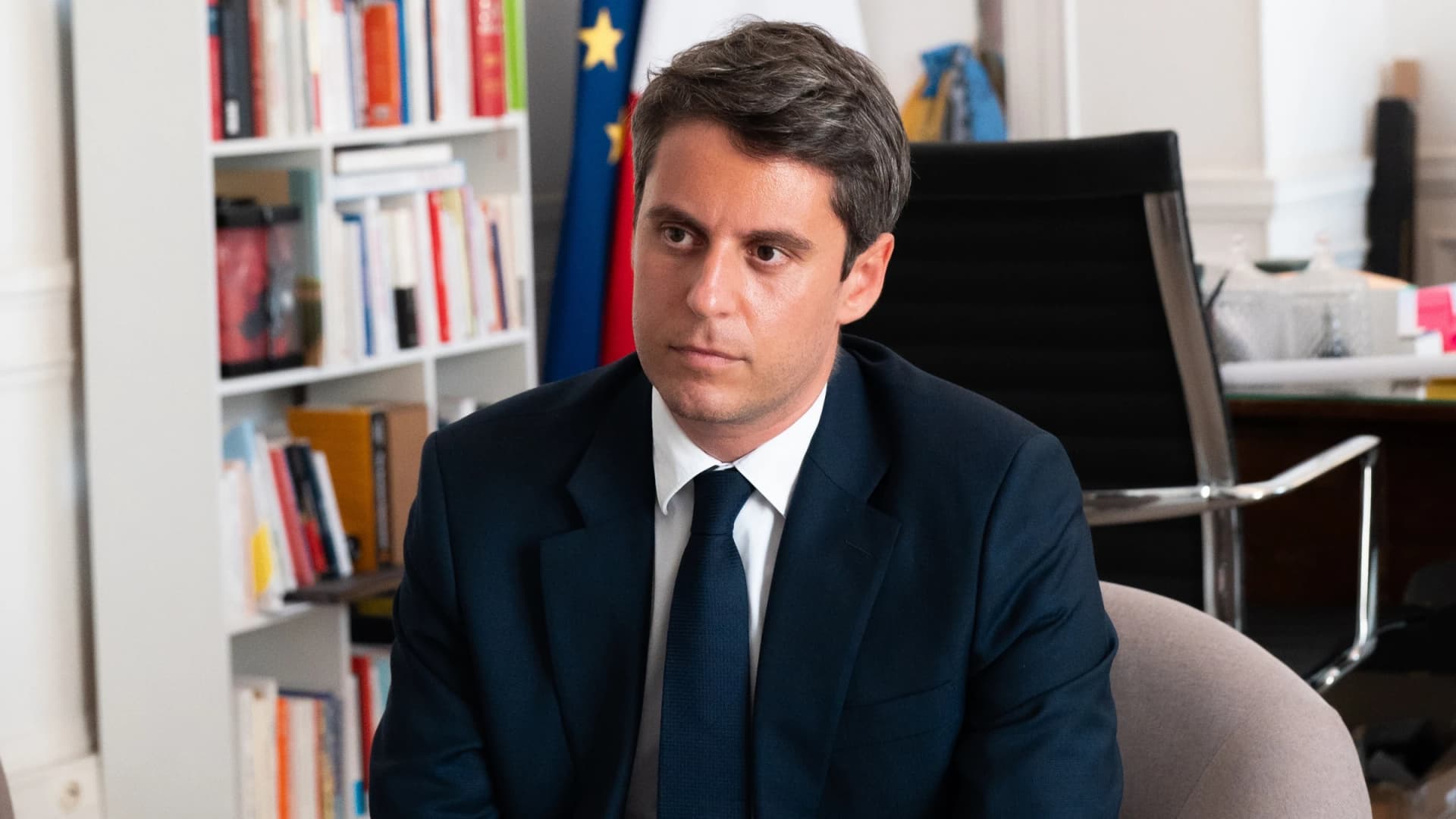 Portrait en plan moyen de Gabriel Attal assis dans un fauteuil gris à l'intérieur d'un bureau officiel. Il porte un costume bleu marine, une chemise blanche et une cravate assortie. À l'arrière-plan, on aperçoit une bibliothèque blanche remplie de livres ainsi que le drapeau européen et le drapeau français.