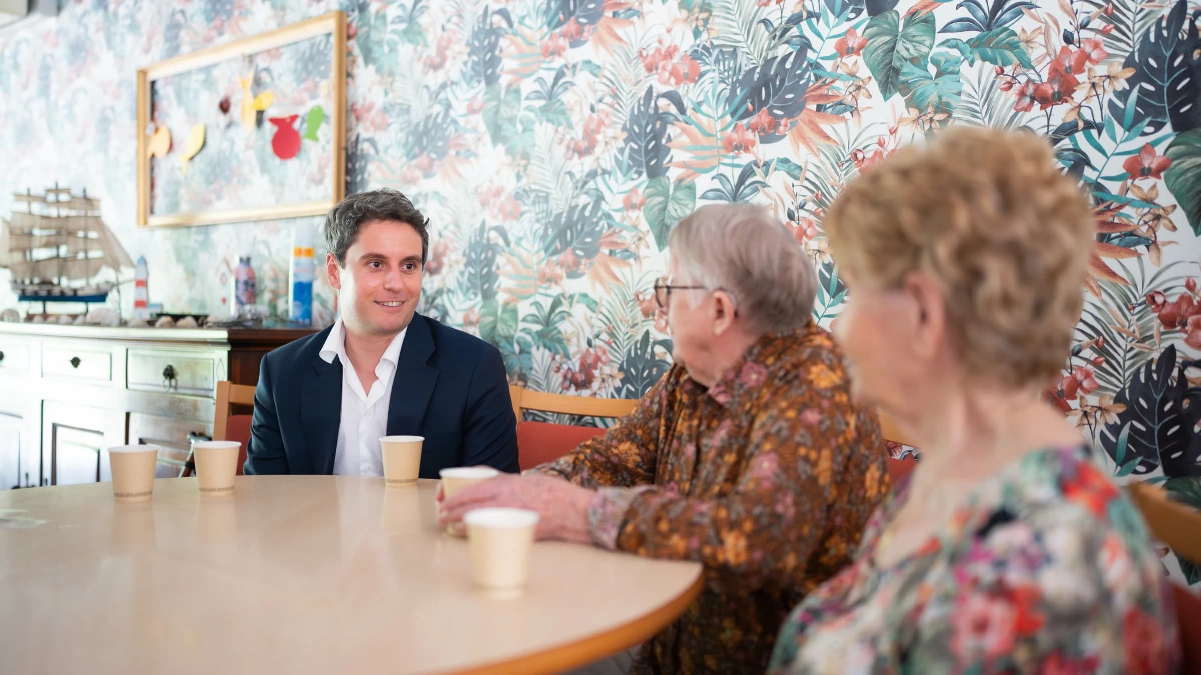 Gabriel Attal en visite dans un EPHAD entouré de deux persones agées souriantes. 