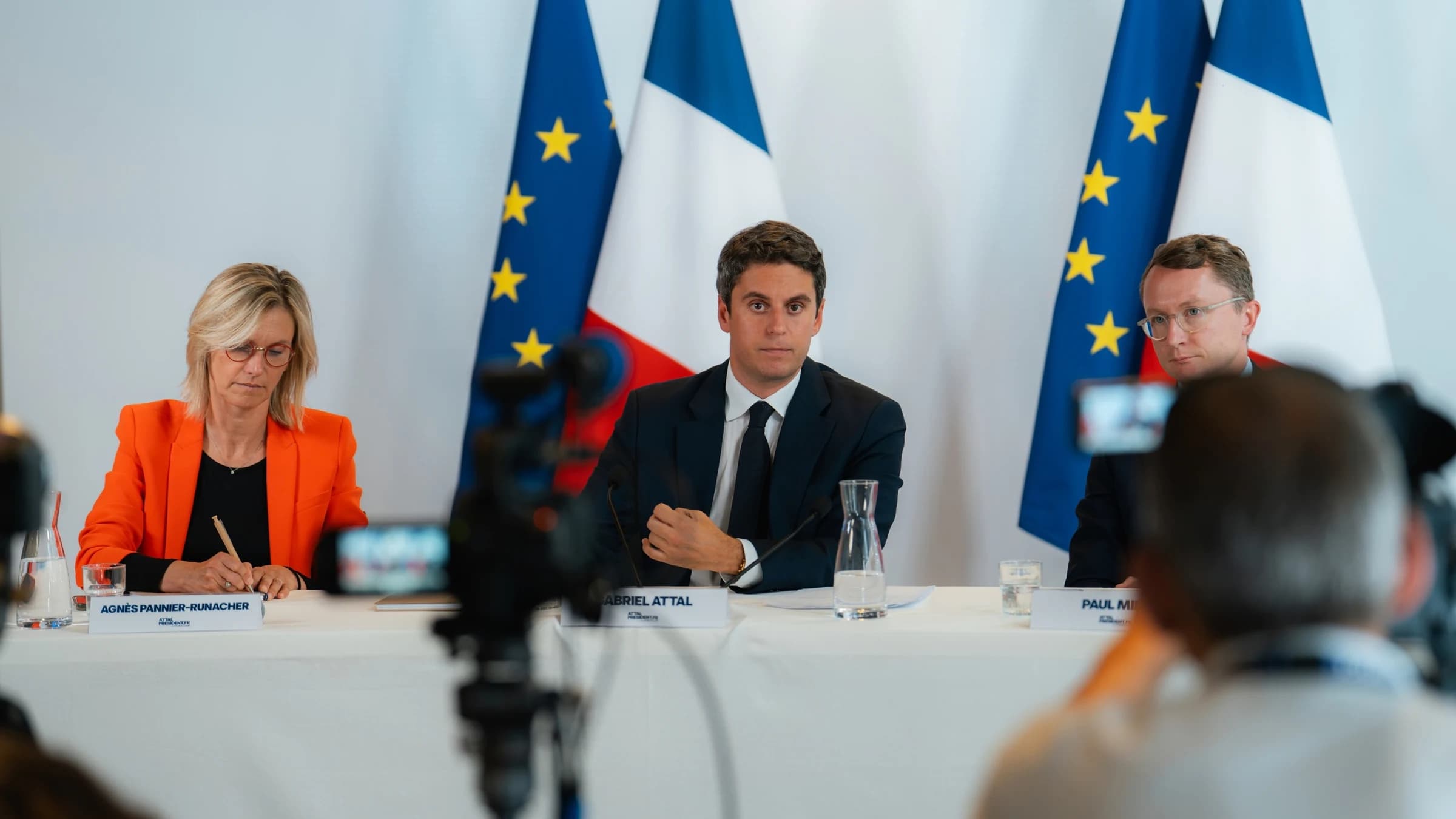 Gabriel Attal, Agnès Pannier Runacher, Paul Midy assis avec en fond des drapeaux français et européens en fond et un air sérieux