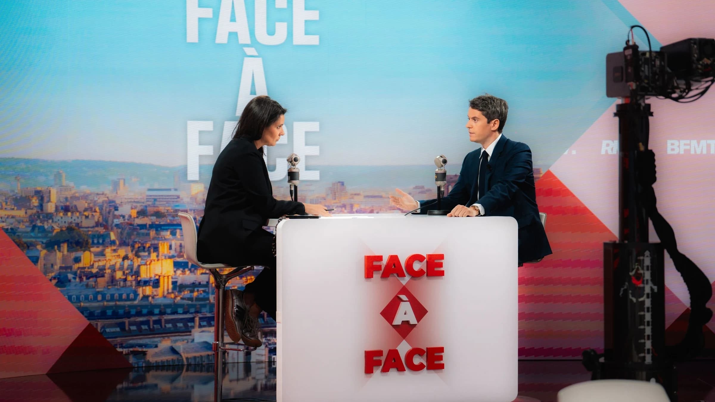 Gabriel Attal sur le plateau BFMTV face à Apolline de Malherbe parlant au micro. 