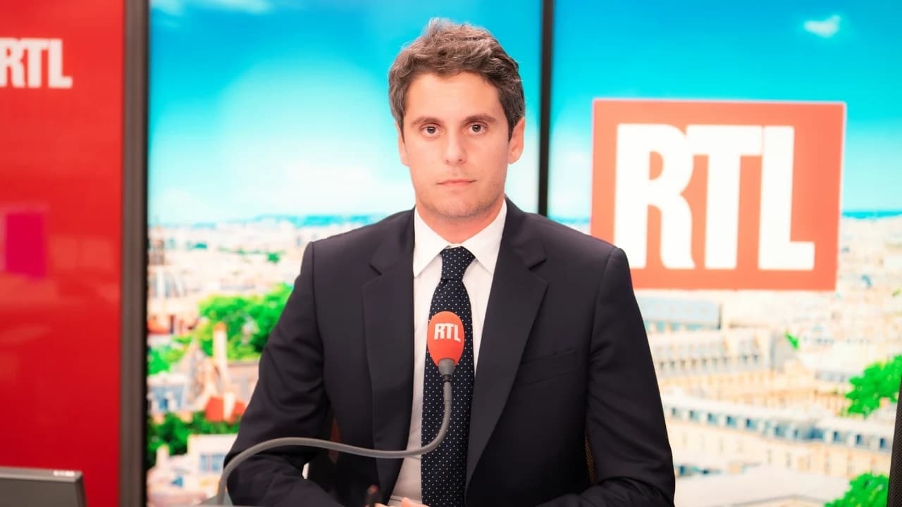 Gabriel Attal au micro de la radio RTL. Il fait face à un microphone marqué du logo RTL, avec en arrière-plan un écran affichant une vue de Paris sous un ciel bleu et le logo rouge et blanc de la station.