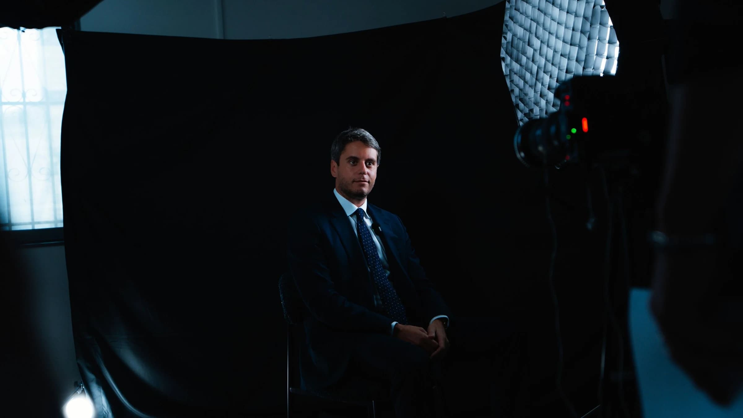 Gabriel Attal sur un fauteuil avec un fond noir souriant d'un air rassurant 