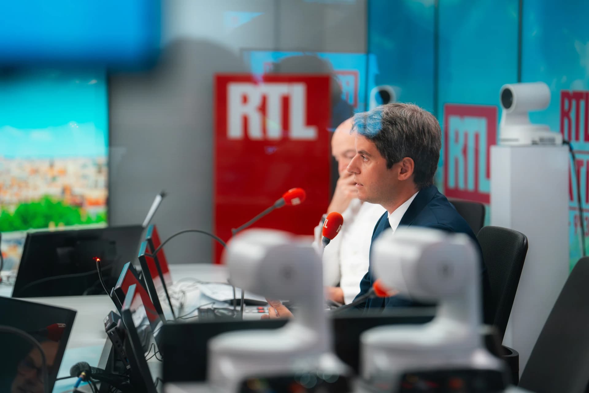 Gabriel Attal de profil dans le studio de RTL, en costume sombre, assis face à un micro rouge de la station, avec le logo RTL en arrière-plan et du matériel de régie au premier plan.