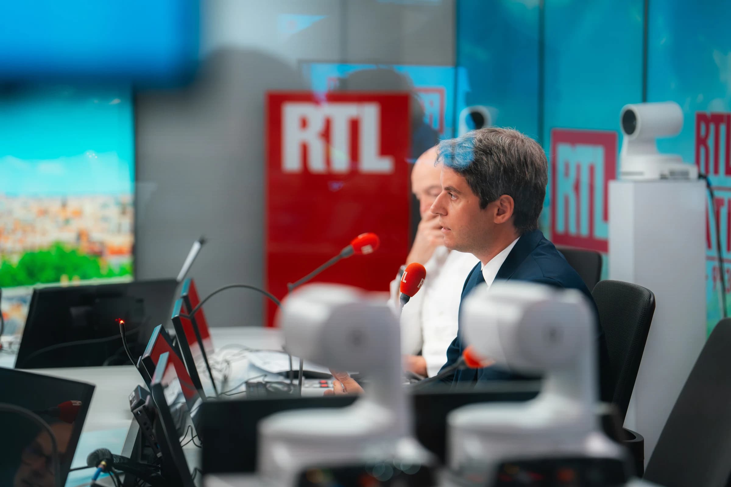 Gabriel Attal de profil dans le studio de RTL, en costume sombre, assis face à un micro rouge de la station, avec le logo RTL en arrière-plan et du matériel de régie au premier plan.