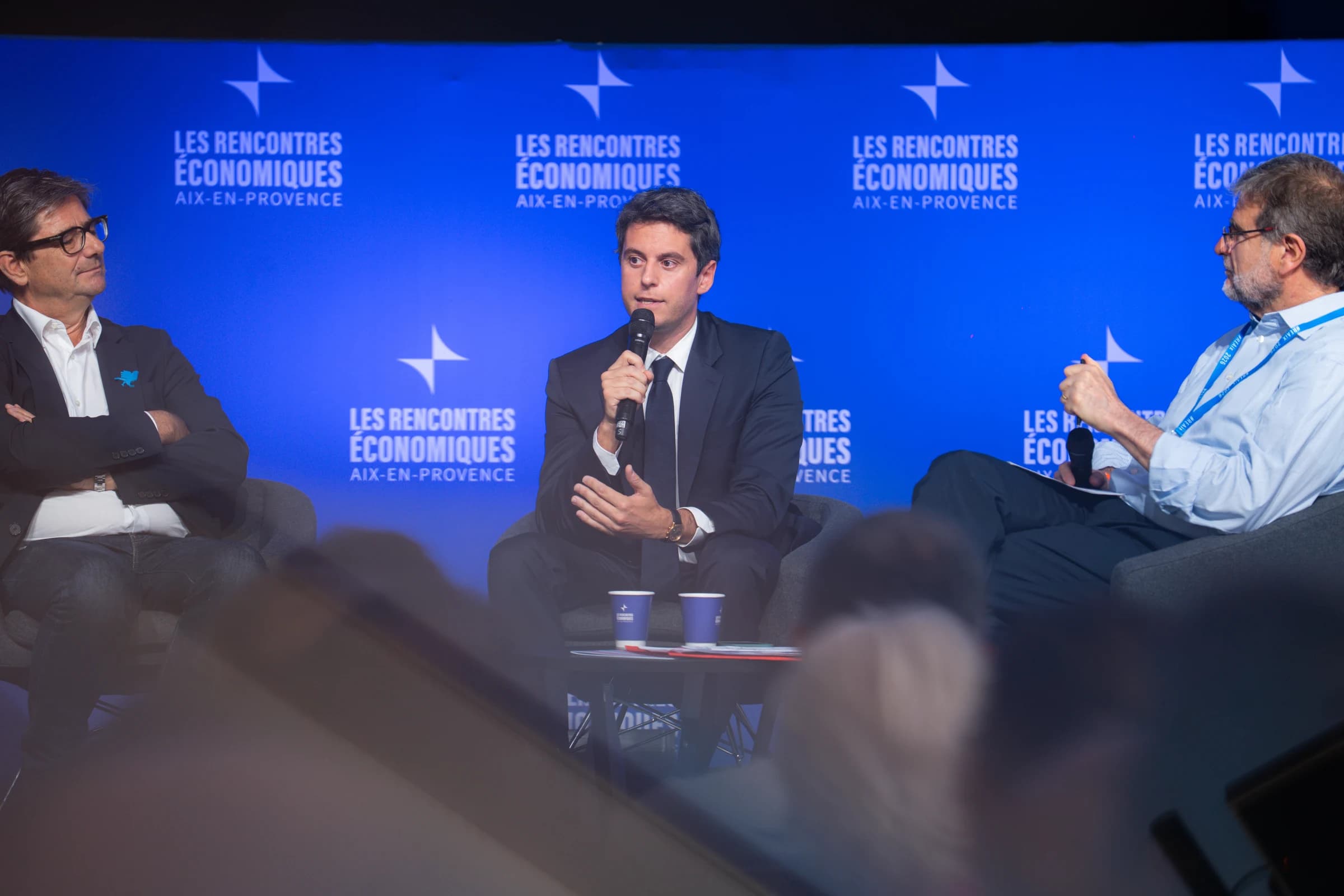 Gabriel Attal s'exprimant au micro lors d'une table ronde aux Rencontres Économiques d'Aix-en-Provence, entouré de deux intervenants sur fond de mur bleu.