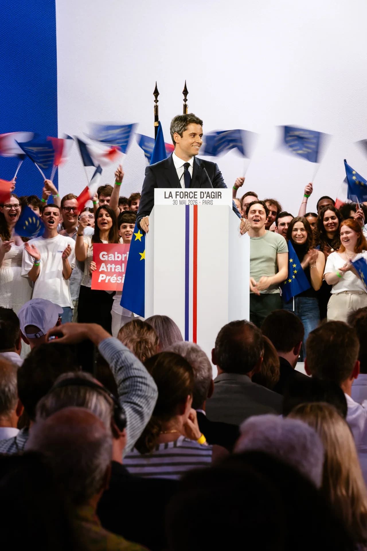 Photographie verticale prise depuis la salle lors du meeting « La Force d'agir » du 30 mai 2026 à Paris. Gabriel Attal, en costume sombre et cravate noire, se tient derrière le pupitre blanc tricolore et sourit. Autour de lui, une foule de jeunes supporters, majoritairement vêtus de blanc, l'acclame avec enthousiasme : certains applaudissent, d'autres agitent des drapeaux français et européens flous par le mouvement, créant un effet de dynamisme. Une pancarte rouge « Gabriel Attal Président ! » est visible à gauche. Au premier plan, le public de dos, debout, regarde la scène. L'atmosphère est chaleureuse et vibrante.