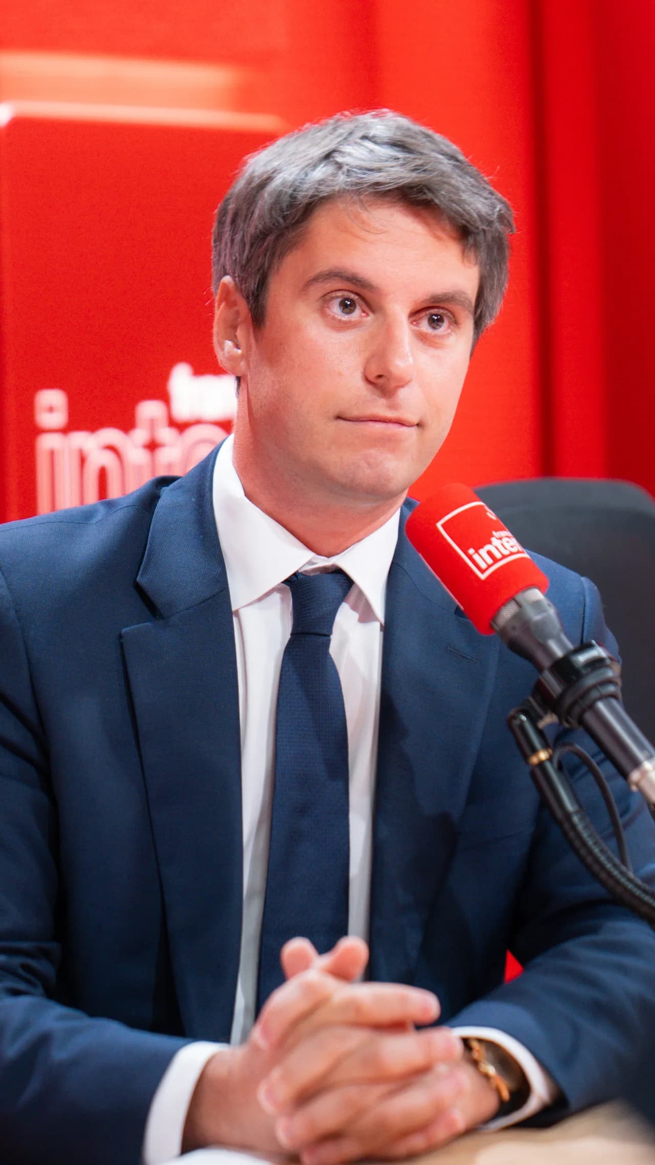 Gabriel Attal en costume bleu marine et cravate, interviewé sur France Inter, mains jointes devant lui, face à un micro rouge de la station.
