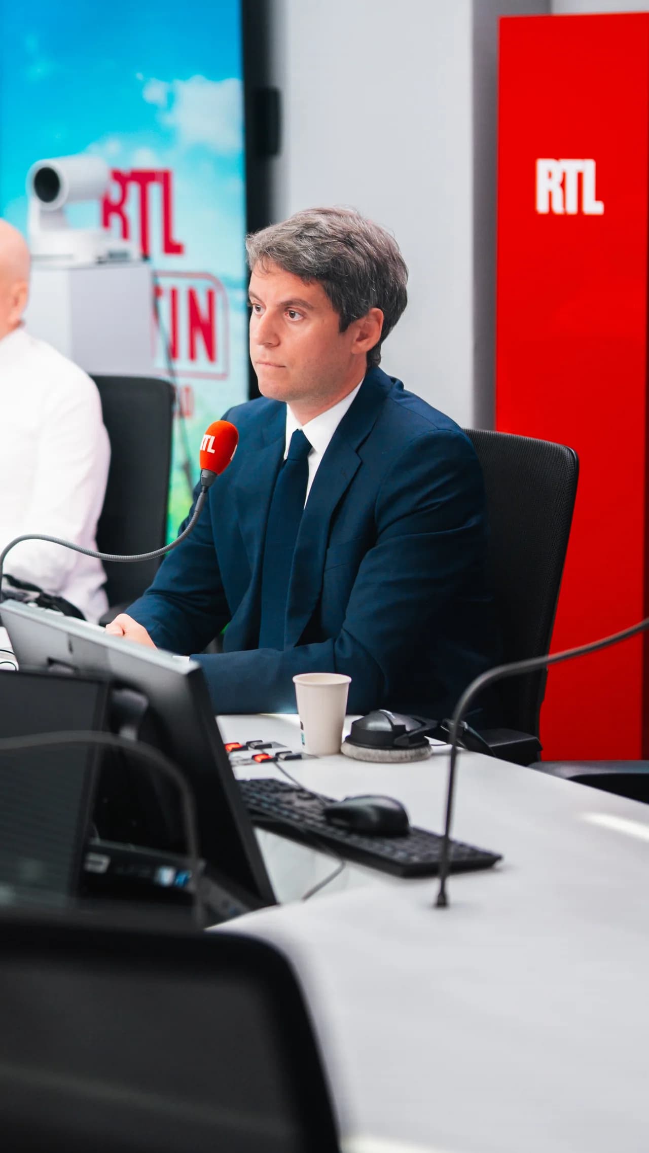 Gabriel Attal de profil dans le studio de RTL, en costume sombre, assis face à un micro rouge de la station, avec le logo RTL en arrière-plan et du matériel de régie au premier plan.