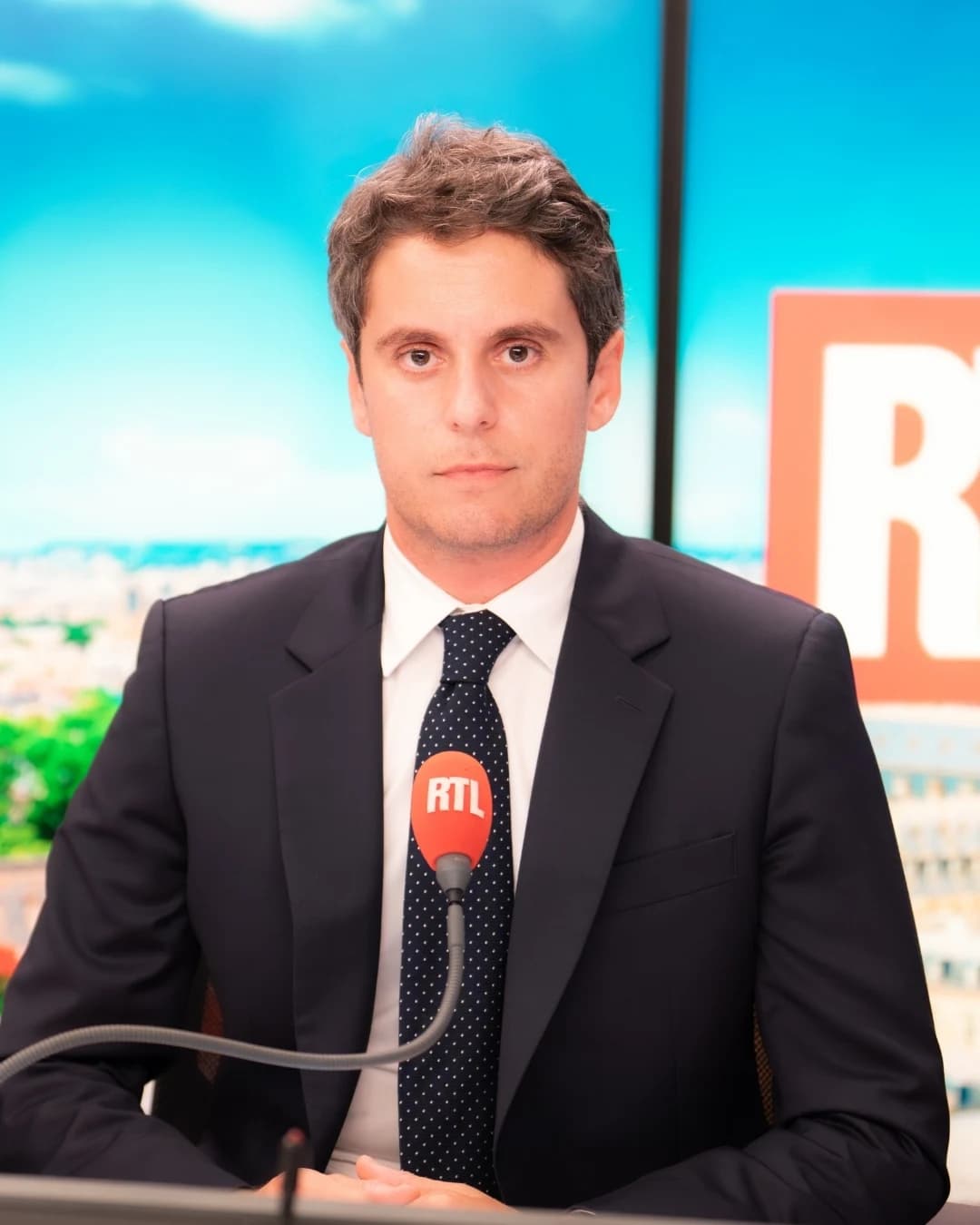 Gabriel Attal au micro de la radio RTL. Il fait face à un microphone marqué du logo RTL, avec en arrière-plan un écran affichant une vue de Paris sous un ciel bleu et le logo rouge et blanc de la station.