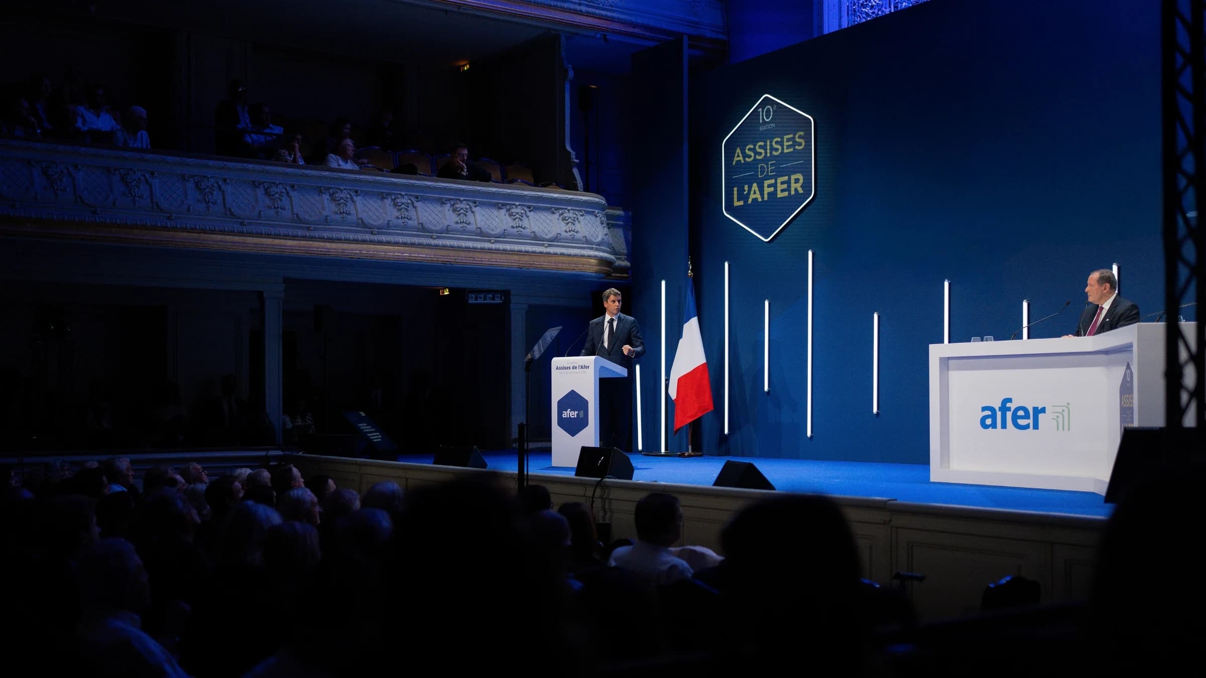 Gabriel Attal aux assises de l'AFER à Paris derrière un pupitre en train de s'exprimeravec en fond des drapeaux français et européens. Il est confiant.