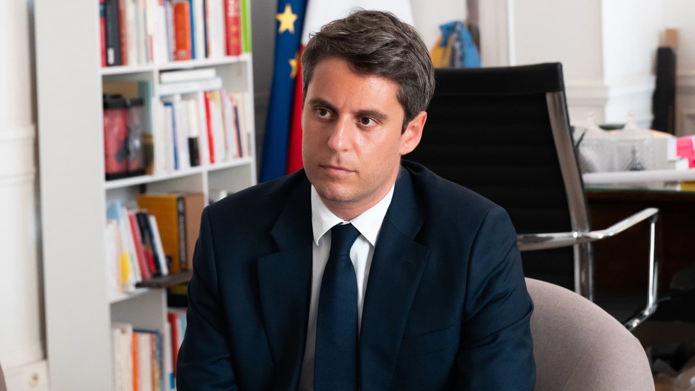 Portrait en plan moyen de Gabriel Attal assis dans un fauteuil gris à l'intérieur d'un bureau officiel. Il porte un costume bleu marine, une chemise blanche et une cravate assortie. À l'arrière-plan, on aperçoit une bibliothèque blanche remplie de livres ainsi que le drapeau européen et le drapeau français.