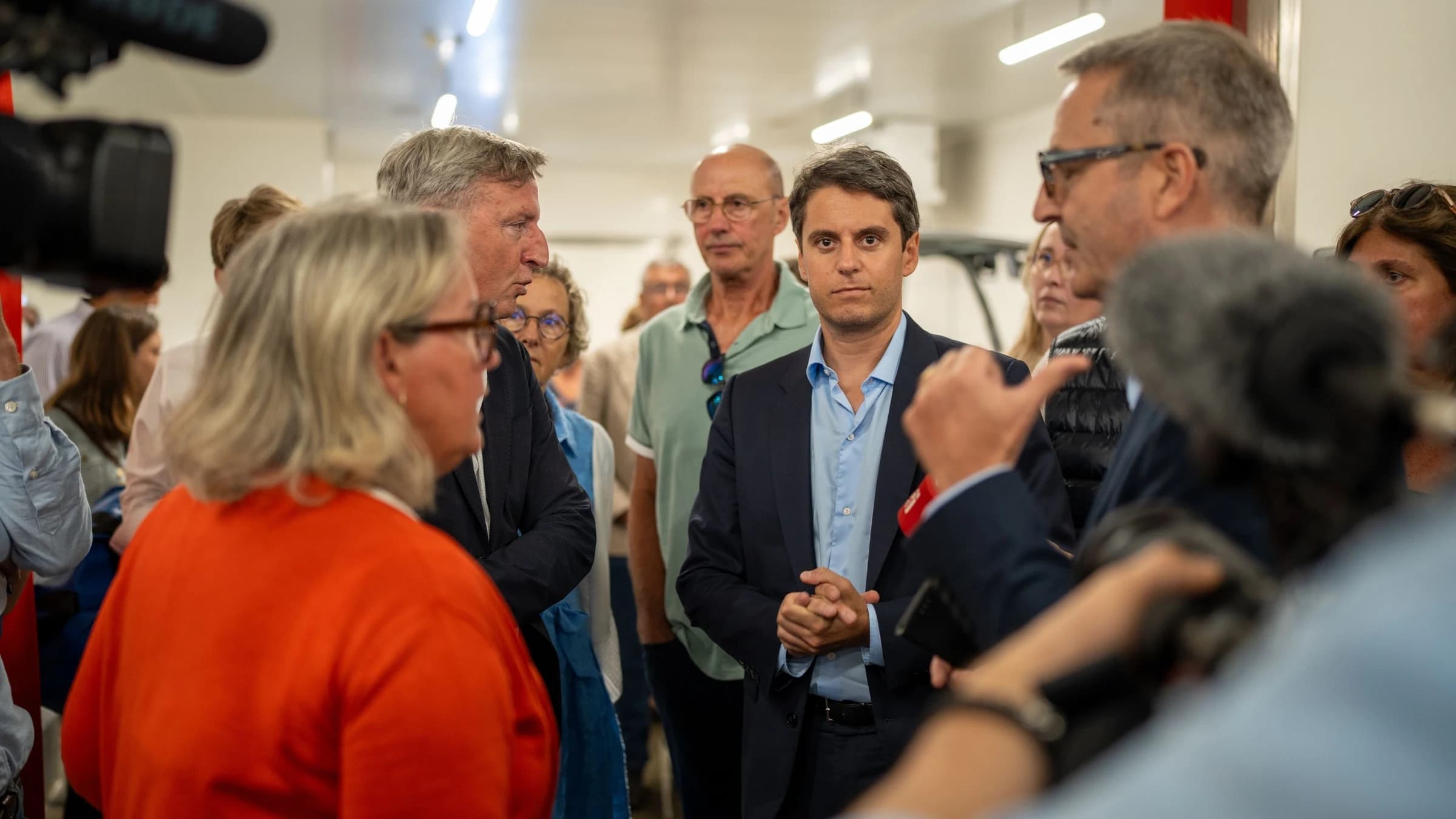 Gabriel Attal en costumen, souriant et etant entouré de beaucoup de personnes dans un lieu lumineux.