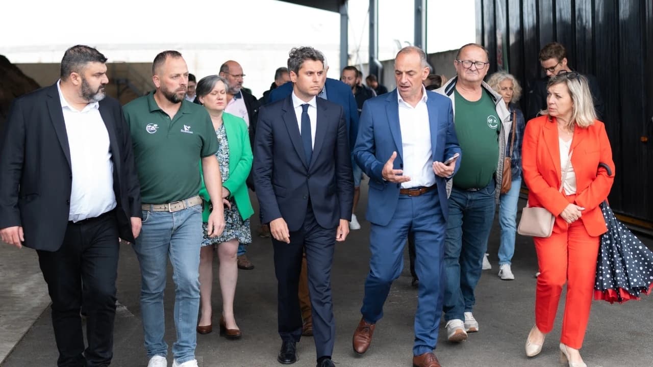 Gabriel Attal marche au centre d’un groupe de personnes dans un bâtiment agricole couvert. Il échange avec un homme en costume bleu, entouré d’élus et d’acteurs locaux
