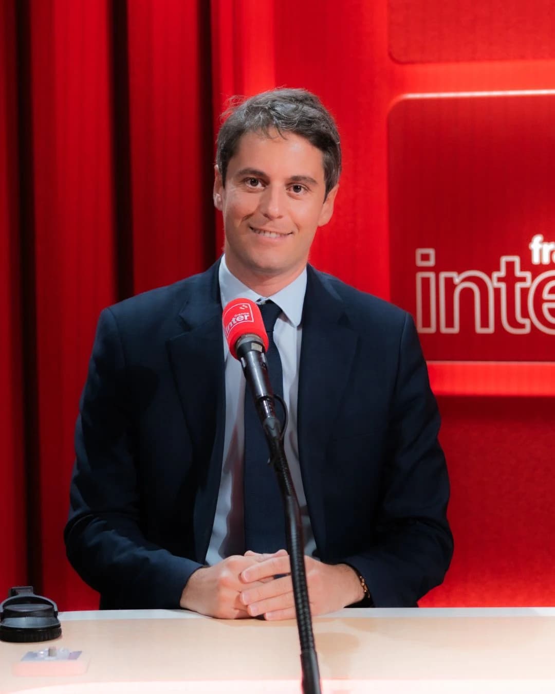 Portrait de Gabriel Attal en costume sombre, assis face caméra à une table de studio devant un micro rouge, avec le logo de France Inter en arrière-plan.