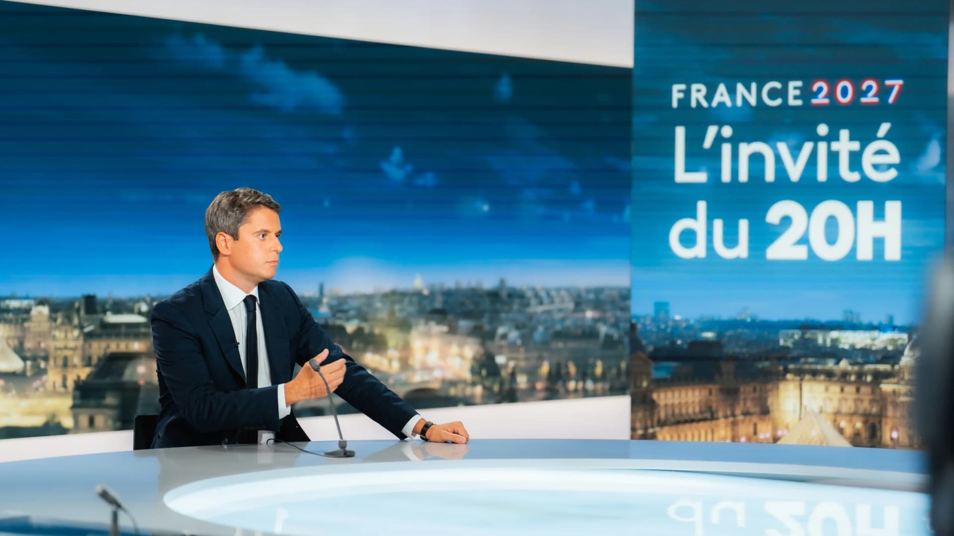 Gabriel Attal en train de s'exprimer derrière un micro en fond il y a le logi du JT de 20H