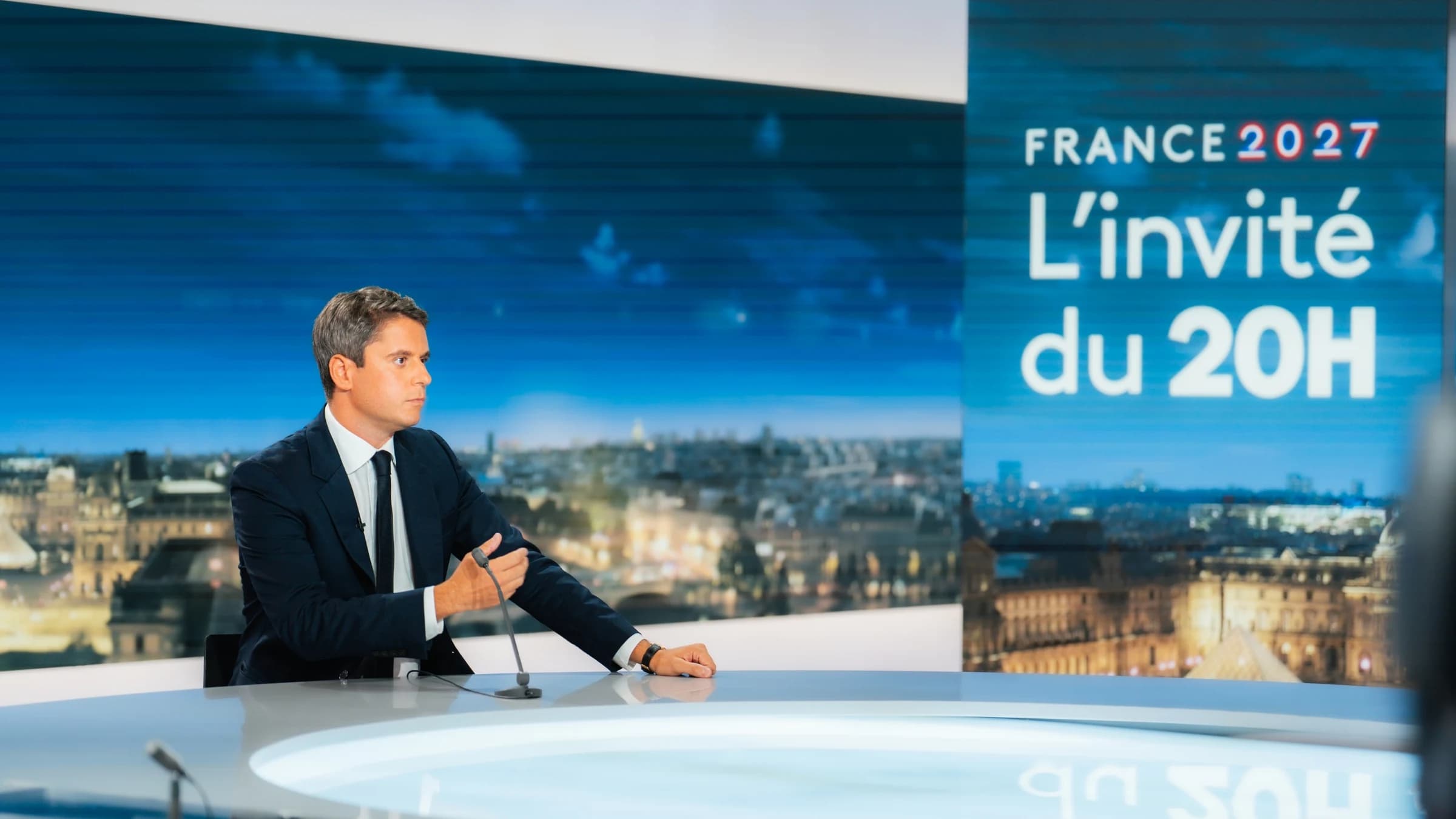 Gabriel Attal en train de s'exprimer derrière un micro en fond il y a le logi du JT de 20H