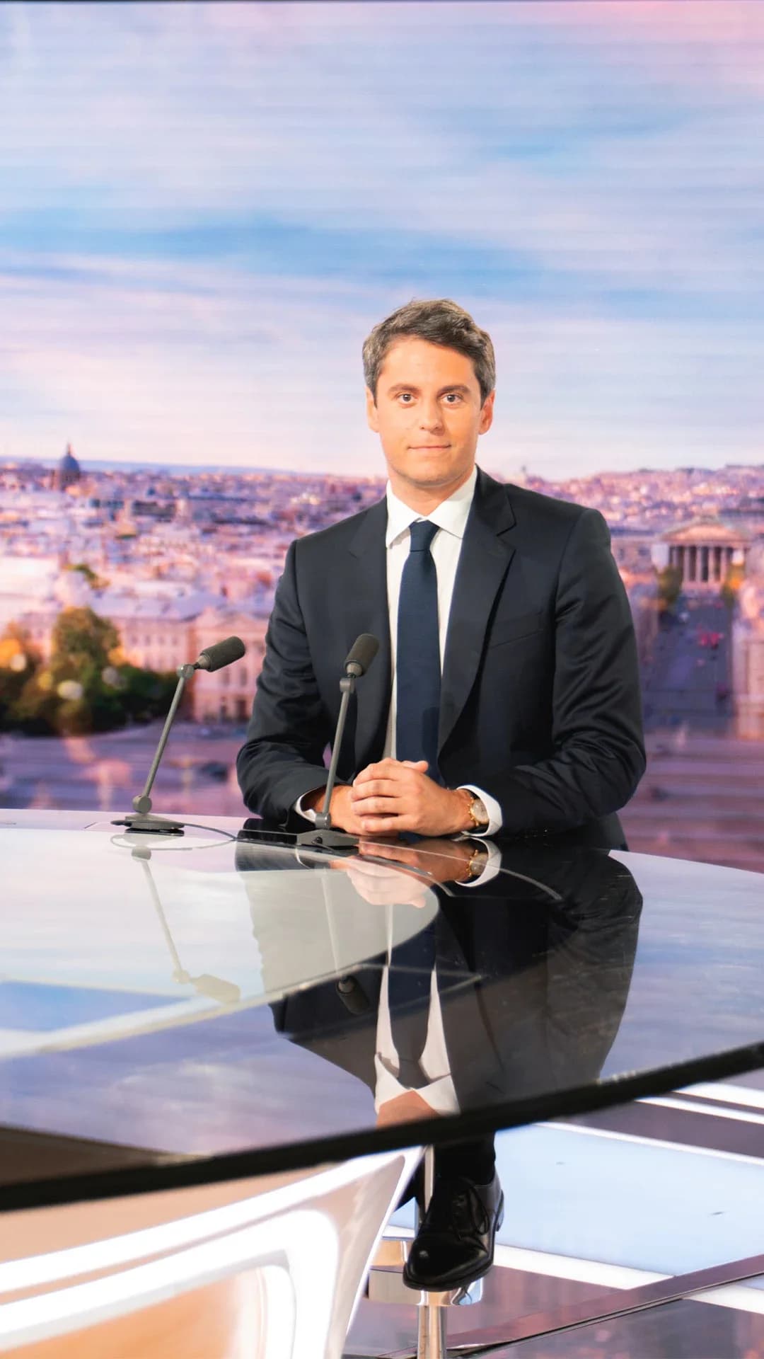 Photographie sur le plateau du journal télévisé de TF1. Gabriel Attal, en costume sombre et cravate bleue, est assis derrière le bureau circulaire réfléchissant du JT, les mains croisées devant lui, regardant face à l'objectif. Deux microphones sont posés sur le bureau. À sa gauche, un grand écran bleu affiche en lettres blanches : « LE 20H – L'INVITÉ – GABRIEL ATTAL ». En arrière-plan, un décor représentant une vue panoramique de Paris aux tons chauds du soir.