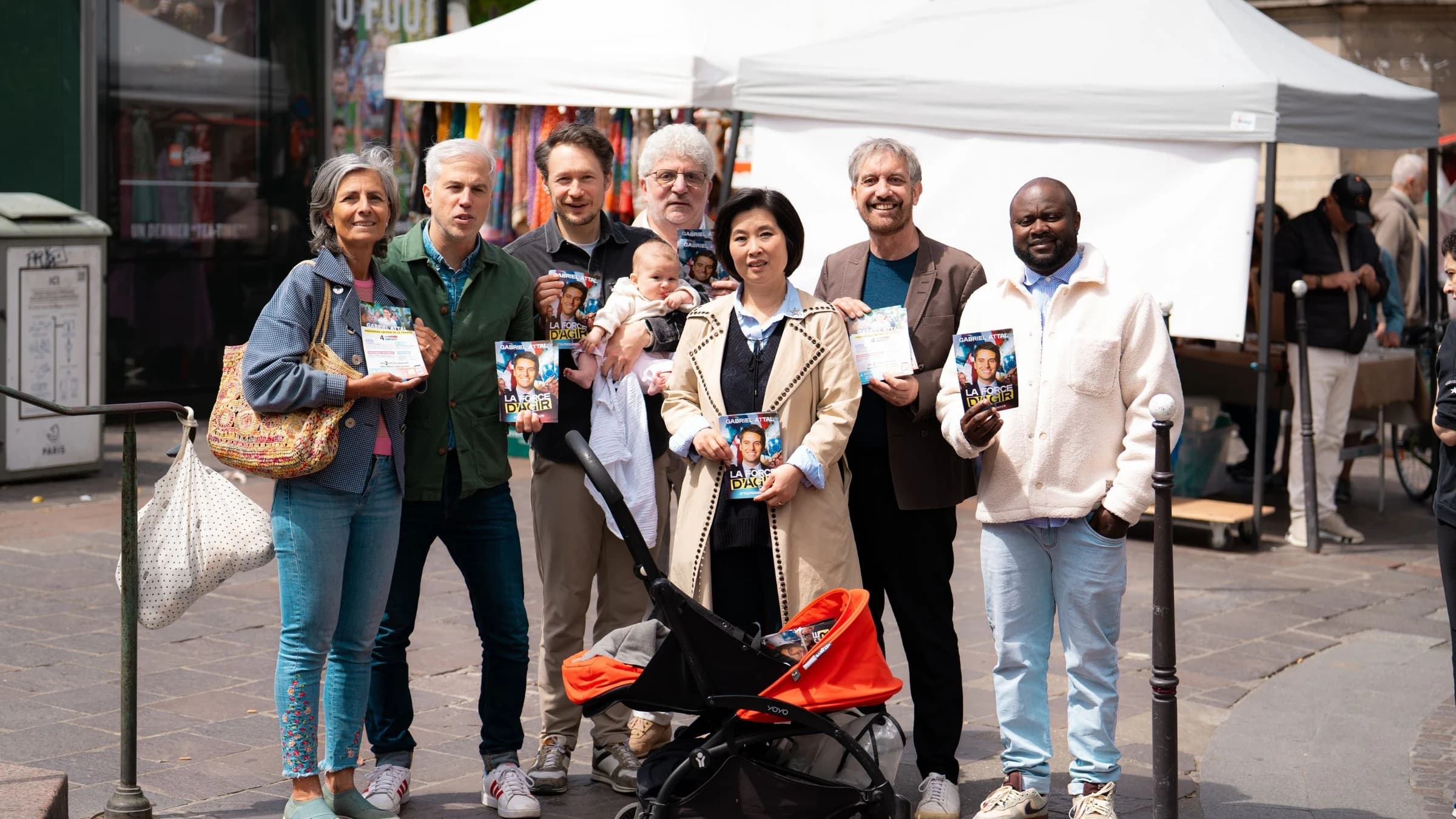 Des militants Renaissance posent sur un marché en tenant des tracts de campagne de Gabriel Attal, dans le cadre d’une opération de mobilisation de terrain.