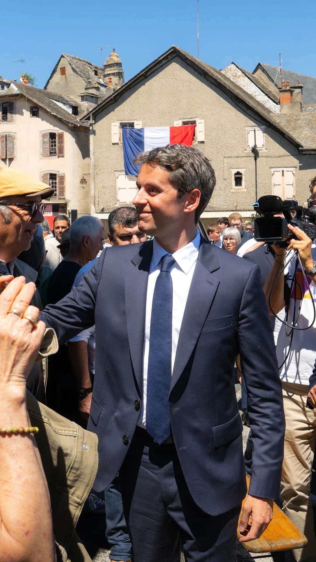 Photographie en extérieur par temps ensoleillé sur une place de village aux bâtisses en pierre grise. Au centre, Gabriel Attal, en costume bleu marine et cravate sombre, avance en souriant au milieu d'une foule. Autour de lui, des habitants l'accueillent chaleureusement — une femme aux cheveux blancs lui tend la main au premier plan — tandis que plusieurs caméramans et photographes captent la scène. À droite, un élu local portant une écharpe tricolore bleu-blanc-rouge l'accompagne. En arrière-plan, un drapeau français est accroché à la façade d'un bâtiment, sous un ciel bleu sans nuages.